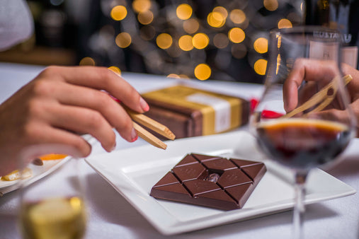 two hands reaching to taste a square chocolate bar set on a white plate