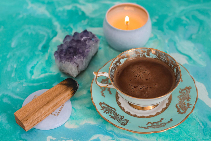 What Exactly Is Ceremonial Cacao? | To'ak Chocolate - image showing a tea cup containing a cacao drink. Beside it is a lit candle, incense, and a purple crystal. Everything is set on a turquoise background
