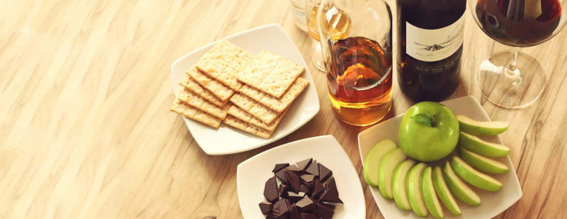 A photograph of a wooden table with bottles of wine and whisky, a glass of wine, a plate of crackers, a plate of sliced green apples, and a plate of To’ak chocolate.