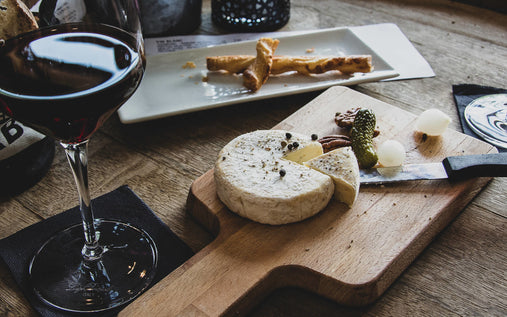 a cheese board sits on a wooden table beside a glass of red wine